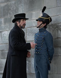 Christian Bale as Augustus Landor Harry Melling as Cadet Edgar Allen Poe meet in the street. Bale wears a long black coat and top hat and Melling is dressed in blue military regalia.