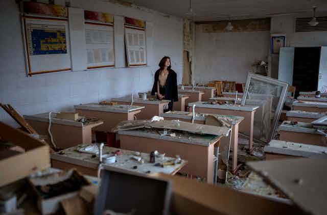 A young woman stands at a table in the middle of a room strewn with rubble.