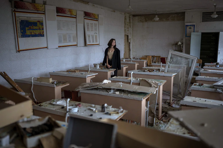 A young woman stands at a table in the middle of a room strewn with rubble.