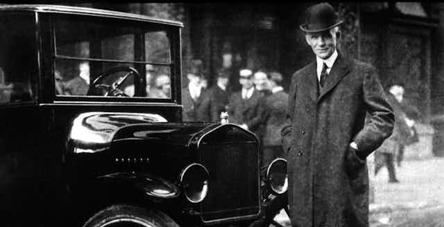 A tall man in a wool coat, tie and formal hat stands alongside an old-timey car.