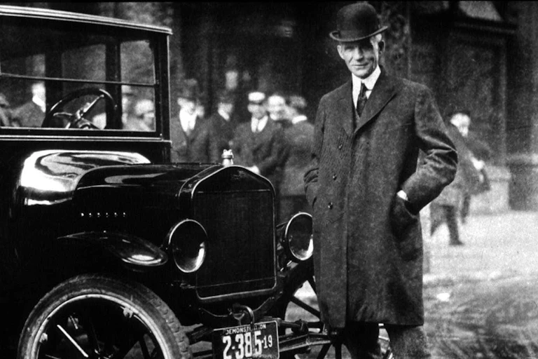 A tall man in a wool coat, tie and formal hat stands alongside an old-timey car.