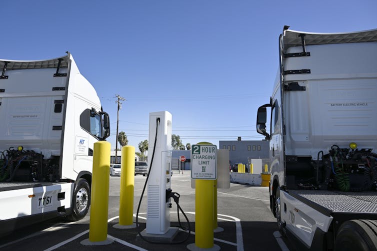 Two electric truck cabs are parked on either side of a charger with a sign reading '2 hour charging limit'.