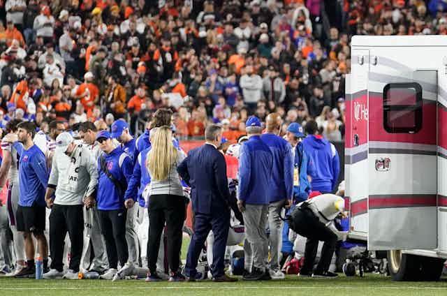 A crowd of people gather on a football field in front of supports in the crowd. An ambulances is also on the field.