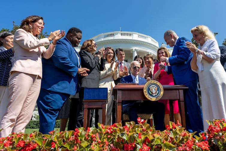President Joe Biden surrounded by a number of people sitting at a desk in front of the White House.