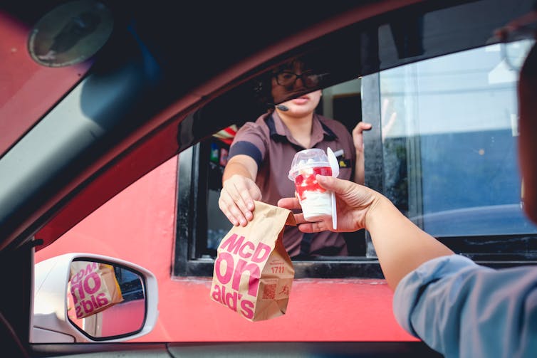 A fast food worker at a drive through window handing a bag of food and a drink to a customer