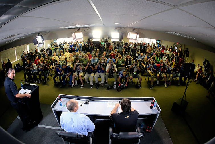 Man stands at podium speaking before a mass of reporters.