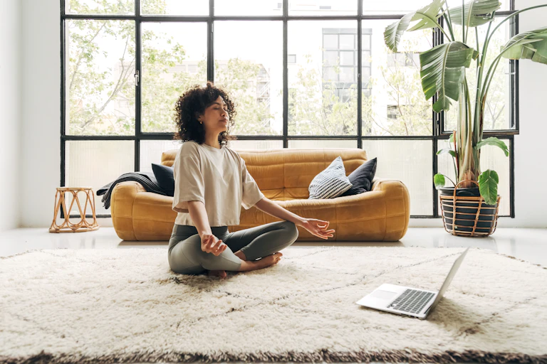 A young multiracial woman sits on a carpeted floor in a crosslegged position with eyes closed, meditating in front of a laptop.