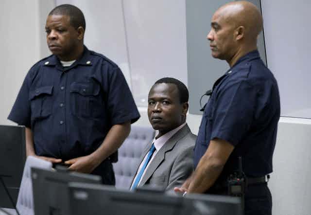 Wearing a gray suit, Dominic Ongwen sits, looking directly at the camera, while two security guards stand on either side of him