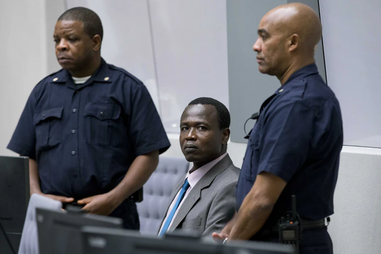 Wearing a gray suit, Dominic Ongwen sits, looking directly at the camera, while two security guards stand on either side of him