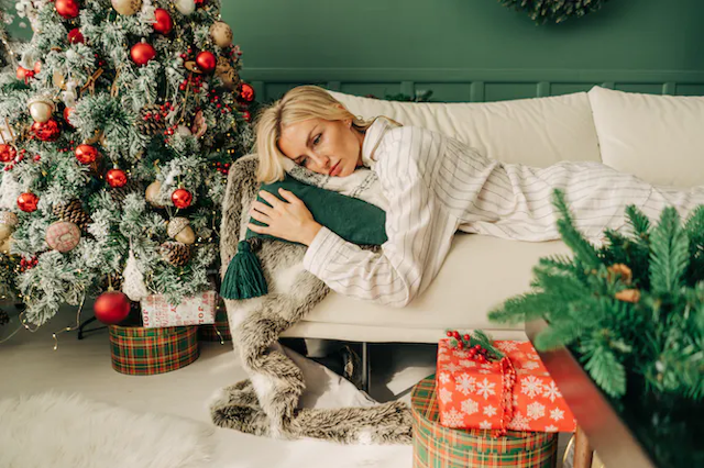Seorang perempuan yang tampak lelah berbaring di sofa di samping pohon Natalnya.