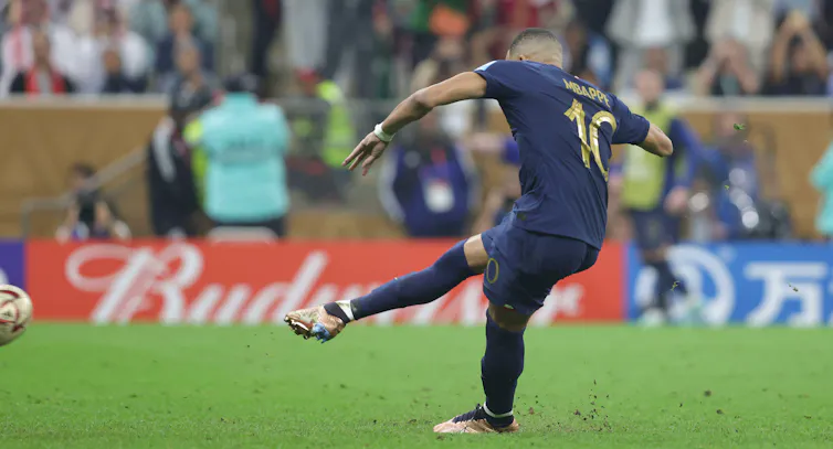A footballer mid-strike in a dark blue kit on a green pitch.