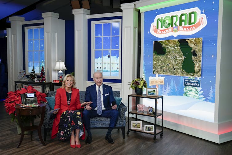 A man and a woman sit next to a large screen that says NORAD Tracks Santa.