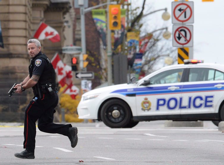 A police officer runs with his gun drawn. A police cruiser and Canadian flags are behind him.