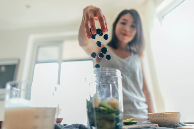 A young woman in her kitchen drops some blueberries into a blender.