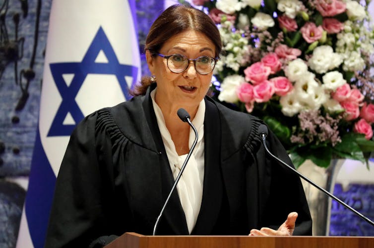 A dark-haired woman with glasses in front of an Israeli flag, talking into some microphones.