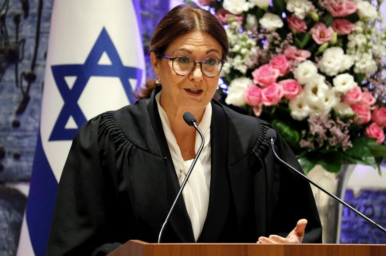 Una mujer de cabello oscuro con anteojos frente a una bandera israelí, hablando por unos micrófonos.