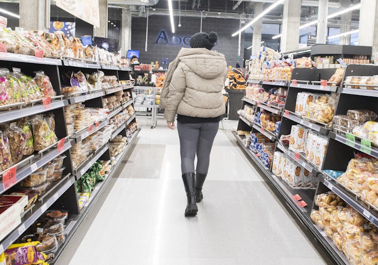 A woman in a parka, jeans and boots walks down a grocery store aisle.