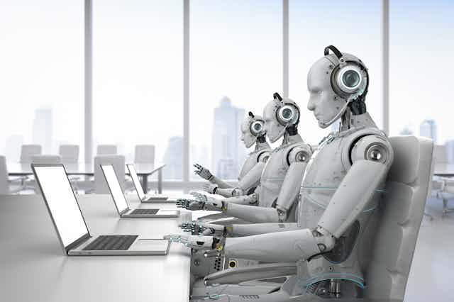 three humanoid robots seated at a desk typing on laptops