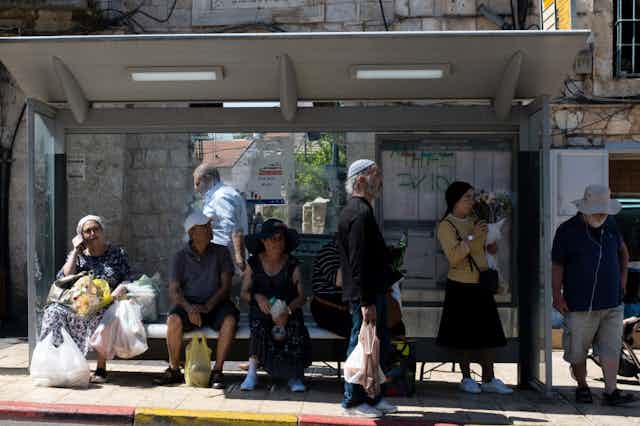 Half a dozen people wait under a shelter at a bus stop.