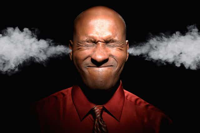 A bald-headed Black man wearing a wine-colored shirt and tie grimaces with his eyes closed and has steam coming out of his ears. The background is black.