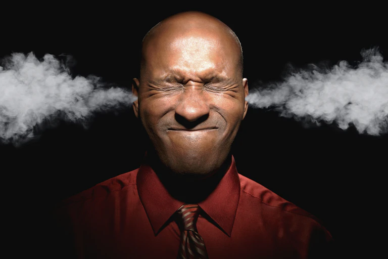 A bald-headed Black man wearing a wine-colored shirt and tie grimaces with his eyes closed and has steam coming out of his ears. The background is black.