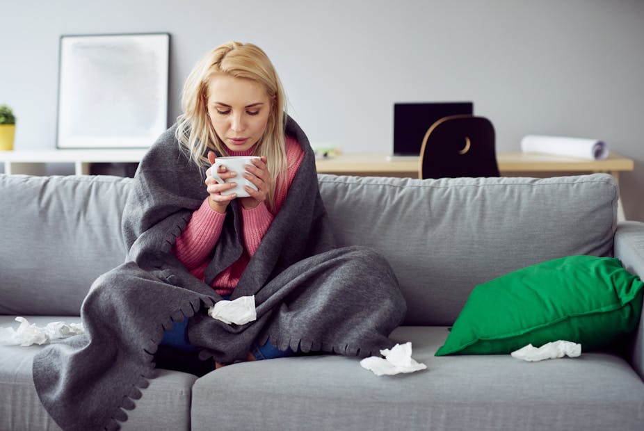 Sick blonde woman sitting on sofa wrapped in a blanket and holding a cup of tea. There are crumpled tissues on the couch.