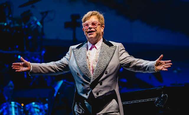 Elton John wears pink sunglasses and a grey double breasted suit on stage. His arms are outstretched.