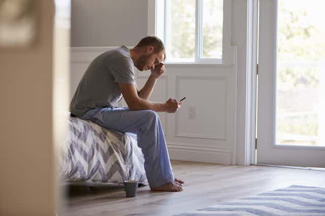 A man sits on the edge of his bed, hunched over his phone.