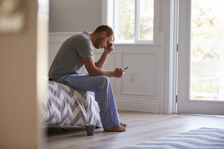 A man sits on the edge of his bed, hunched over his phone.