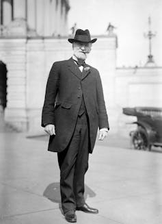 Una foto en blanco y negro muestra a un hombre de pie con un vestido de época de 1900.