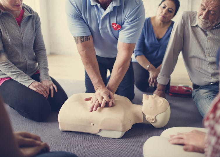 A person in a blue shirt with hands on the chest of a dummy, while other people observe.