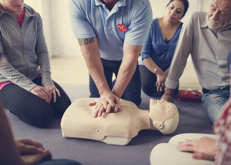 A person in a blue shirt with hands on the chest of a dummy, while other people observe.