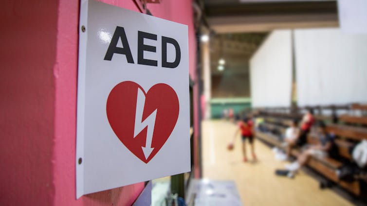 A sign reading AED with a heart icon in the foreground, with athletes in a gymnasium in the background.