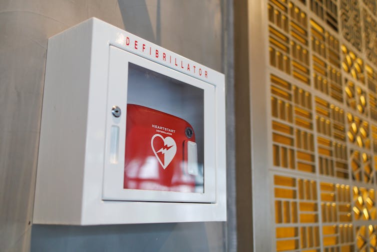 A white box reading Defibrillator with a red box inside it hanging on a wall