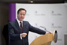 A dark-haired man in a suit stands behind a podium, gesturing as he speaks.