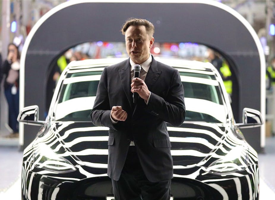 Elon Musk in a black suit speaking into a microphone in front of a black Tesla, people in the background.