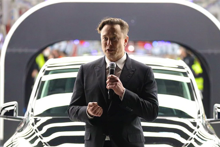 Elon Musk in a black suit speaking into a microphone in front of a black Tesla, people in the background.