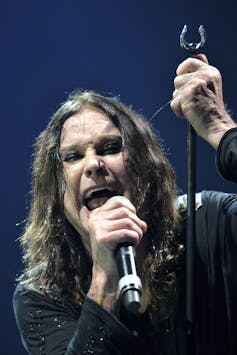 Ozzy Osbourne performs, singing into a microphone with long black hair and a strained expression.