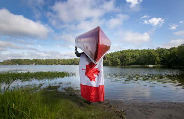 A man carries a canoe into a lake with a Canadian flag on his back.