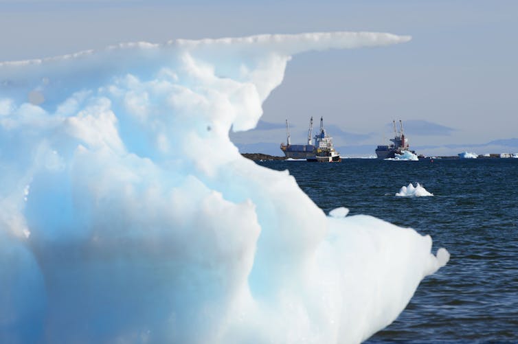 Ships are seen on open water behind a large chunk of blue-ish sea ice.
