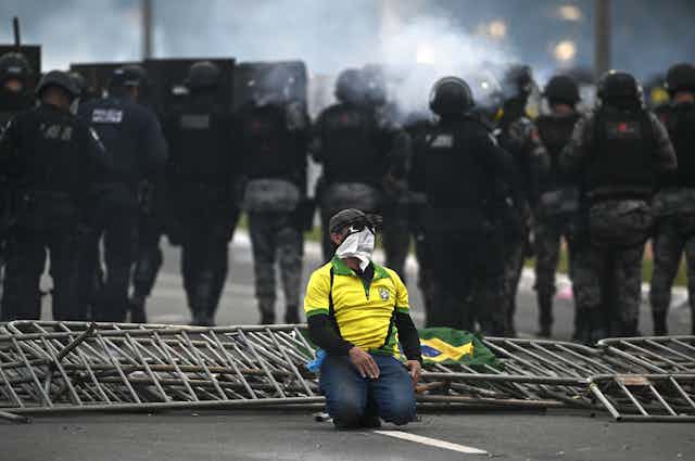 A supporter of ousted Brazilianpresident Jair Bolsonaro wearing the national colours kneels in the street as police parade behind.