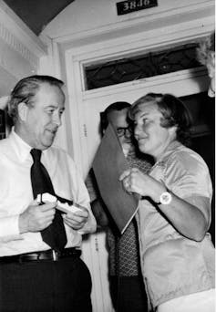 A man in a tie and a woman holding a folder speak to each other.