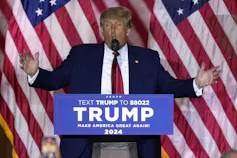A man with orange-blonde hair speaks with his arms spread behind a podium that says Trump. American flags are behind him.