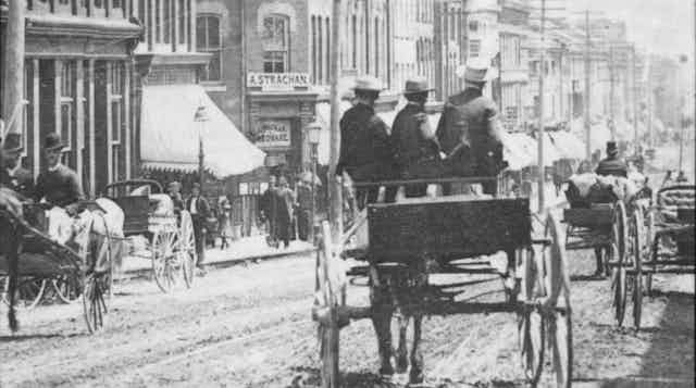 The backs of figures seen riding in a horse-drawn cart on an unpaved bustling city centre street.
