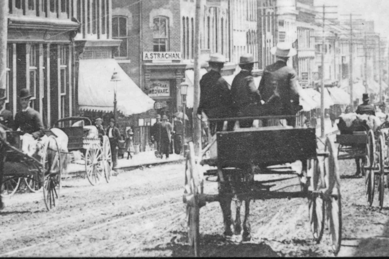 The backs of figures seen riding in a horse-drawn cart on an unpaved bustling city centre street.