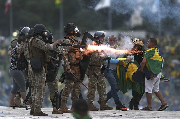 Military police carrying riot gear and firing weapon, next to men wearing Brazilian flags.