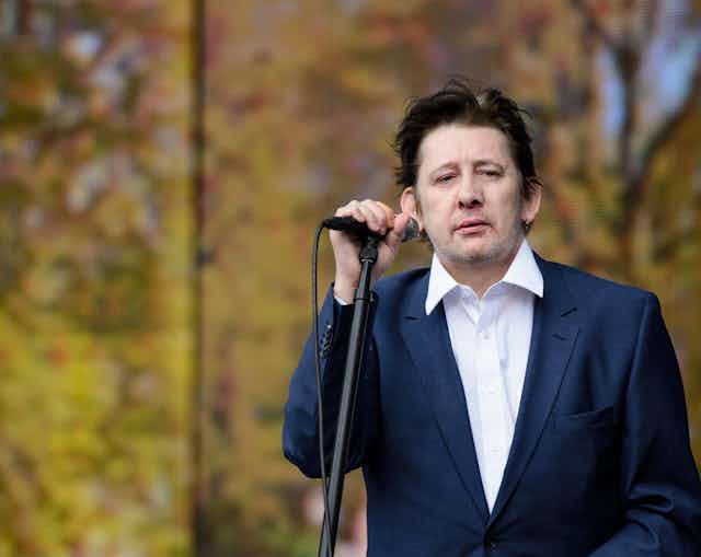Shane MacGowan on stage holding a microphone in a loose fitting blue suit and white shirt.