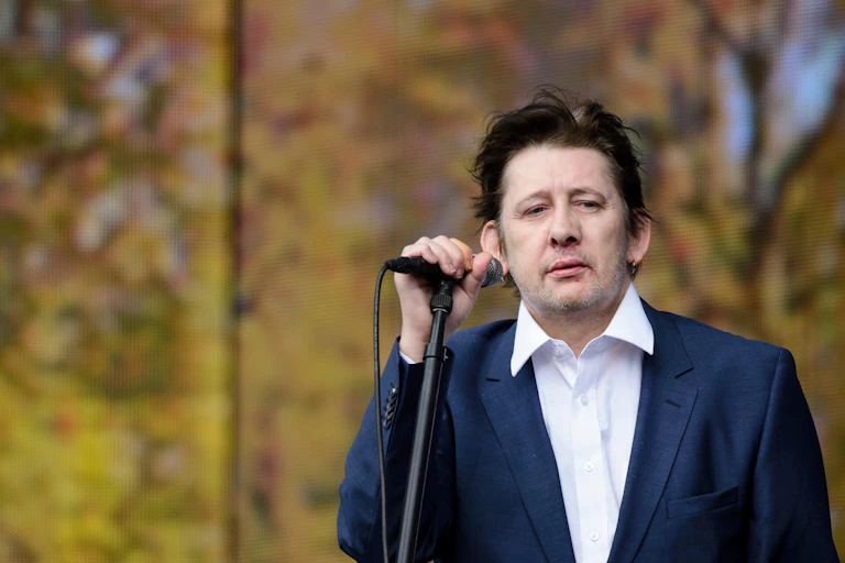 Shane MacGowan on stage holding a microphone in a loose fitting blue suit and white shirt.