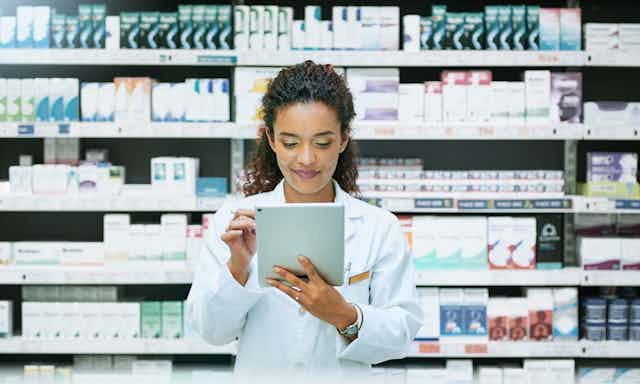 With shelves full of various medications behind her, a young female pharmacist uses a digital tablet while working behind the counter.