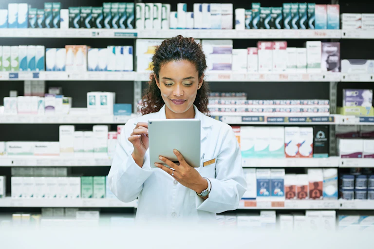 With shelves full of various medications behind her, a young female pharmacist uses a digital tablet while working behind the counter.